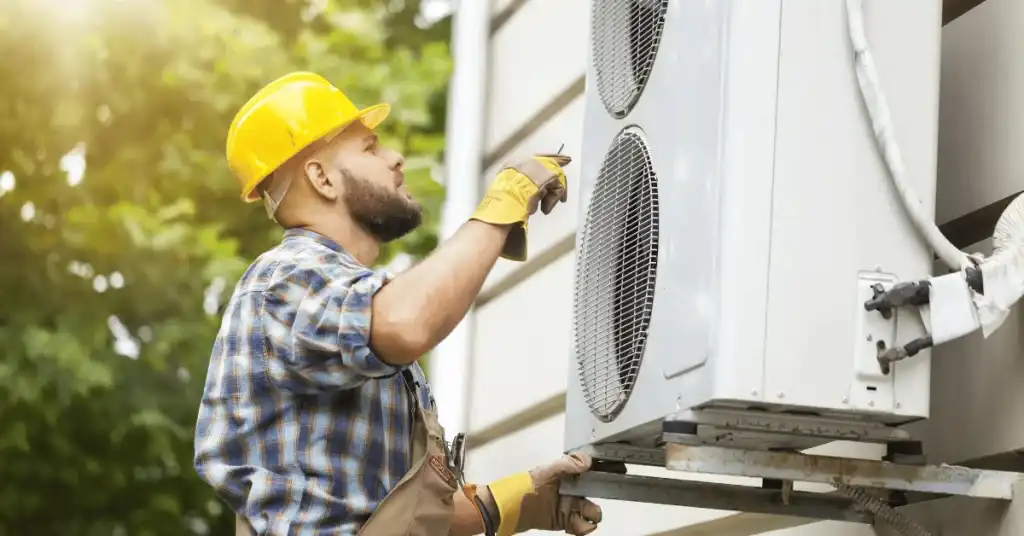 A technician in a yellow hard hat and gloves repairs an outdoor air conditioning unit attached to a wall, showcasing the high-quality AC Repair and Maintenance services by The Chill Brothers in Conroe, TX.